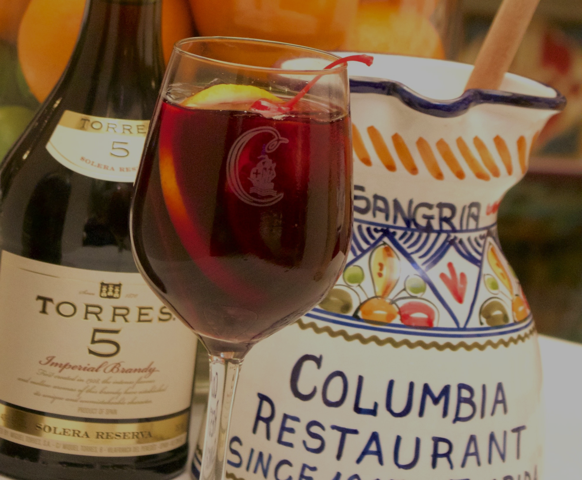 Glass of red sangria next to a bottle of Torres brandy and a decorative sangria pitcher.