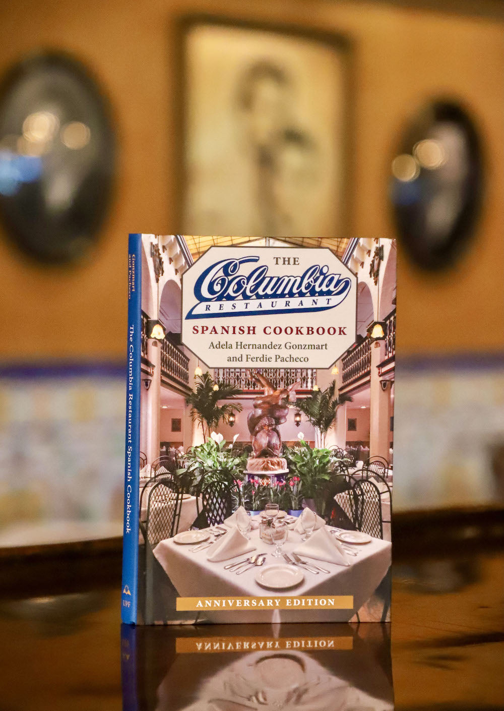 The image shows the cover of the 'Columbia Spanish Cookbook' with a foreword by Andrea Gonzmart Williams. The book is placed on a table with a restaurant setting in the background.